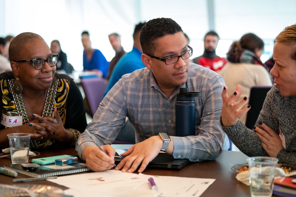 A man sits between two women at a table. They're talking to him as he looks at the woman to his left. He's holding pencil to paper.