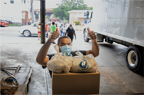A man standing behind a stack of boxes and bags of food, wearing a mask and holding two thumbs up. A truck and other food bank volunteers in the background.