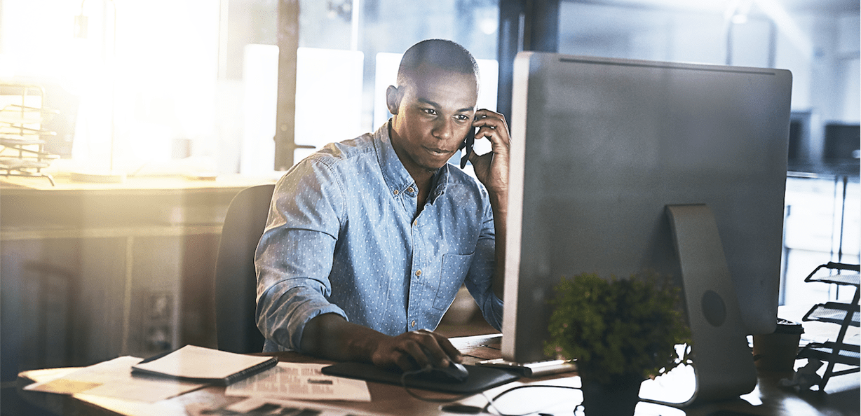 A man sitting at a desk in front of a computer. He is holding a cell phone up to his ear.
