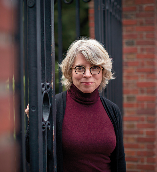 A photo of Jill Lepore, a woman with chin-length light colored hair wearing glasses and a red turtleneck. She stands in a gateway by a brick wall