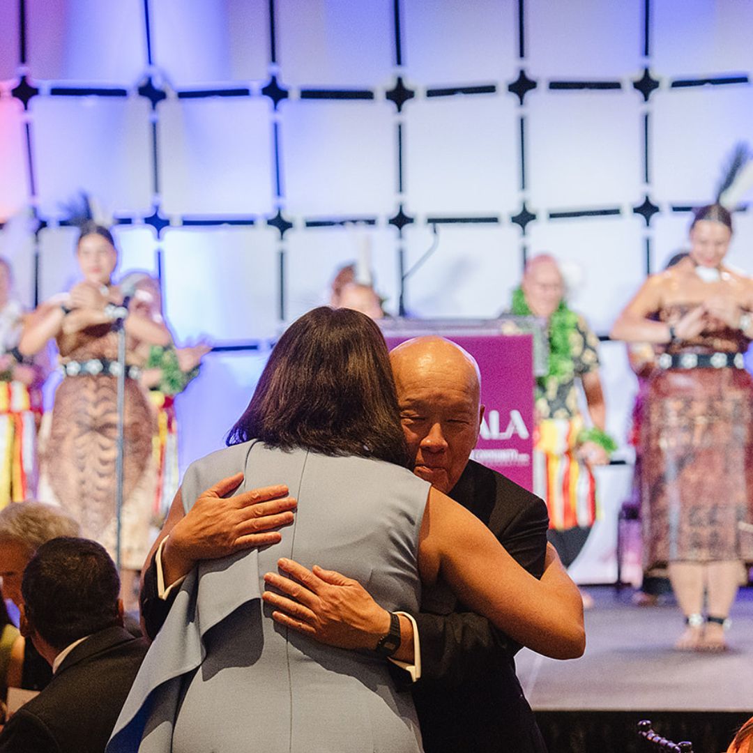 A man and woman embrace at the ACF 2025 gala, behind them dancers perform onstage.