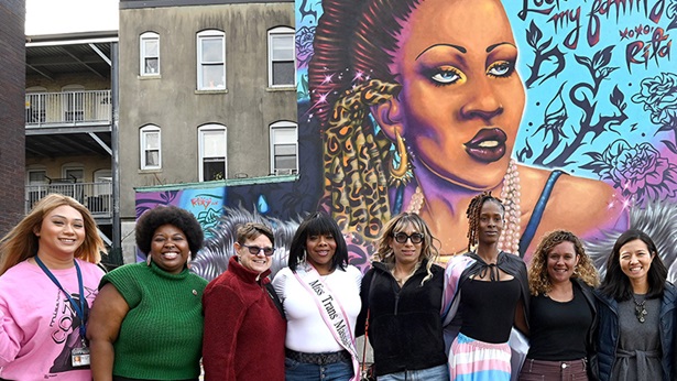 A group of people stands outside in front of a building, on the building is a large colorful memorial mural of a trans woman.
