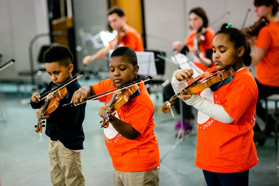 3 young children playing violins in a room with other youth behind them playing violins. They're all wearing orange t shirts.