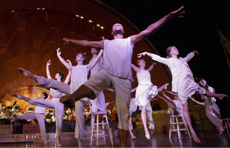 A low-angle photo of dancers wearing loose white clothing, dancing onstage in a triangle formation. Their arms and one of each of their legs are in the ai.r