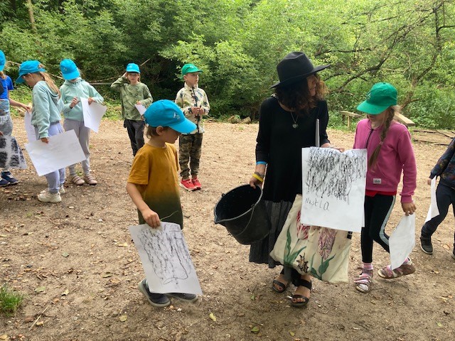 A woman stands with a group of children outside in a clearing, they are holding big pieces of paper and all the children wear matching hats.