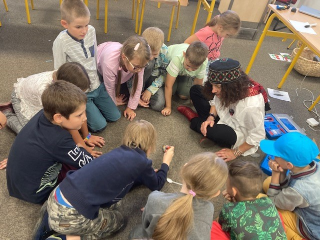 A group of children sit forming a circle on the floor with a woman, they all lean forward looking at an item in the center of the circle.