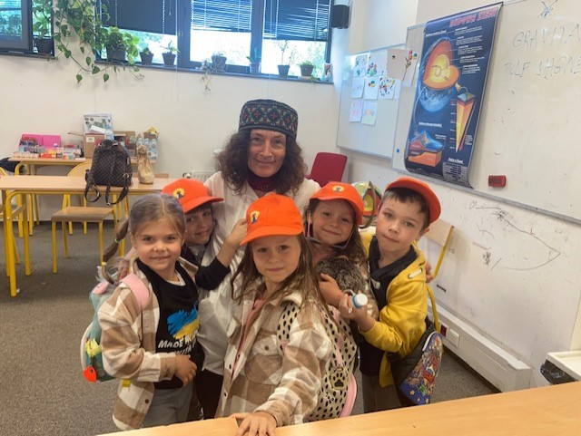 A woman crouches down for a photo with a group of children, they're in a classroom with tables and a whiteboard behind them