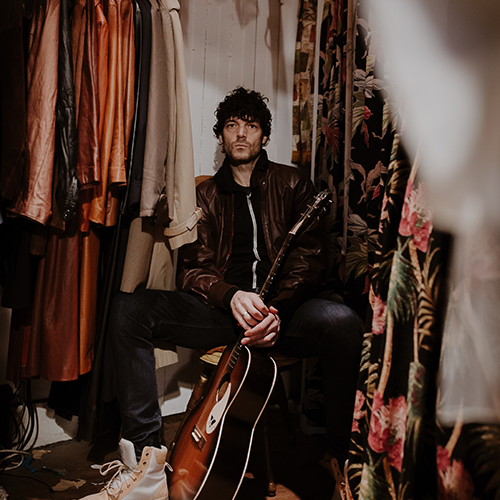 Will Dailey sits in a closet surrounded by racks of clothing, he is holding a guitar