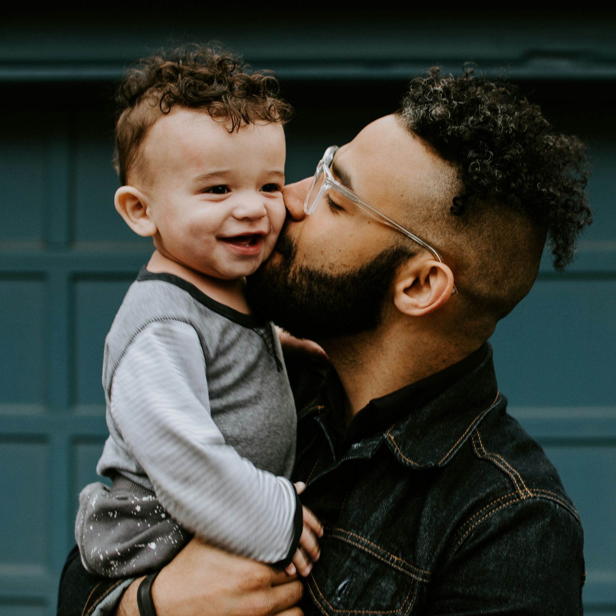 A father, a Latino man, holds his young son and kisses him on the cheek as the child smiles in his father's arms.