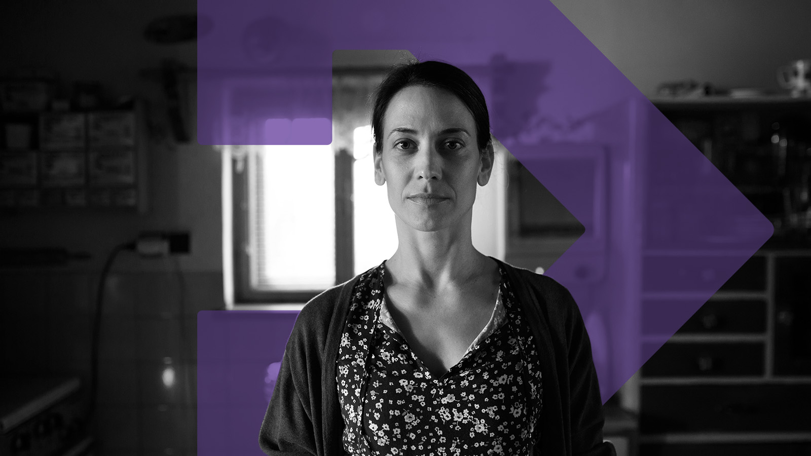 Black and white photo: A woman with very short dark hair stands in a kitchen, the purple TBF arrow is edited into the photo behind the woman.