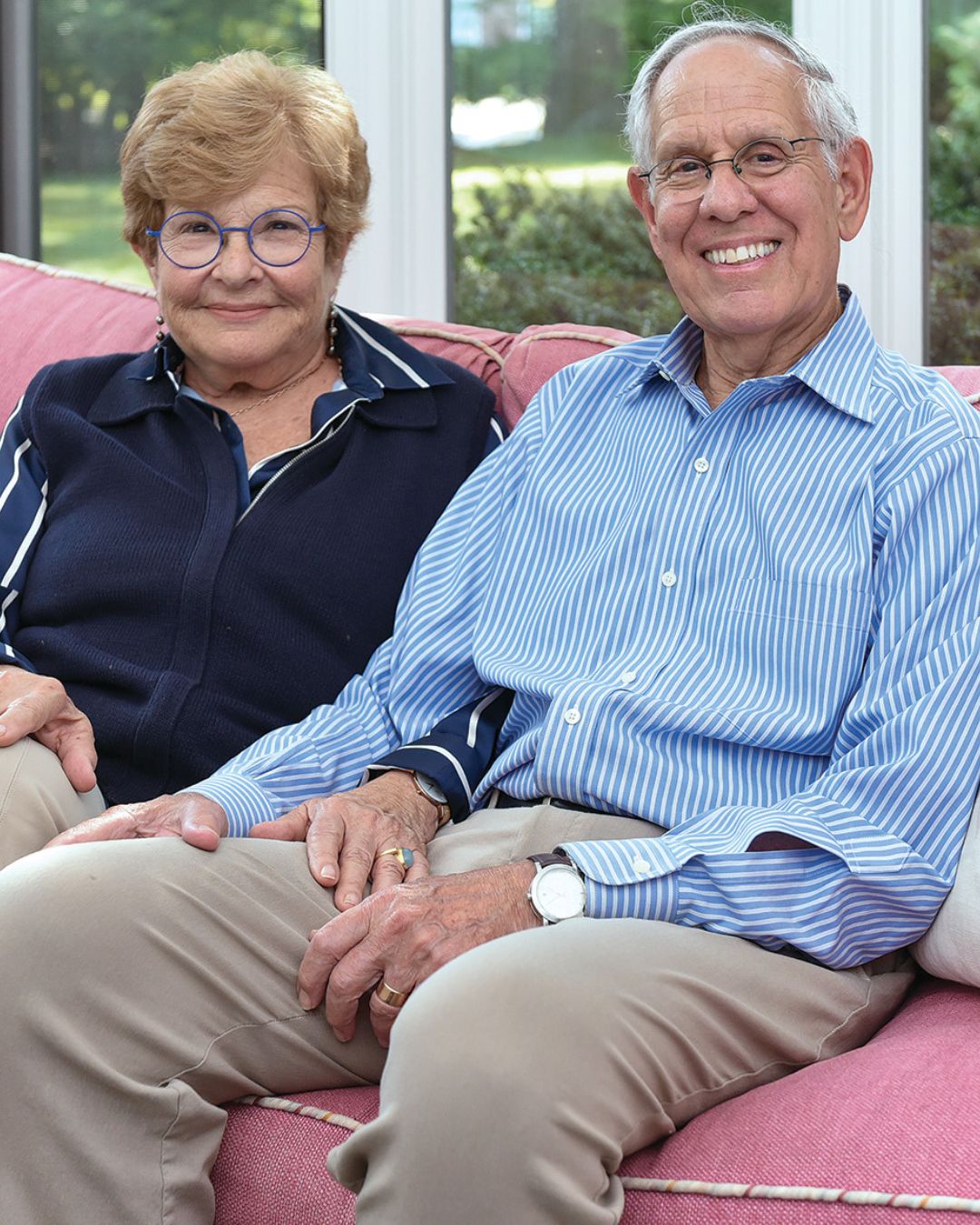 David and Jill Adler sit next to each other on a pink couch in front of large windows