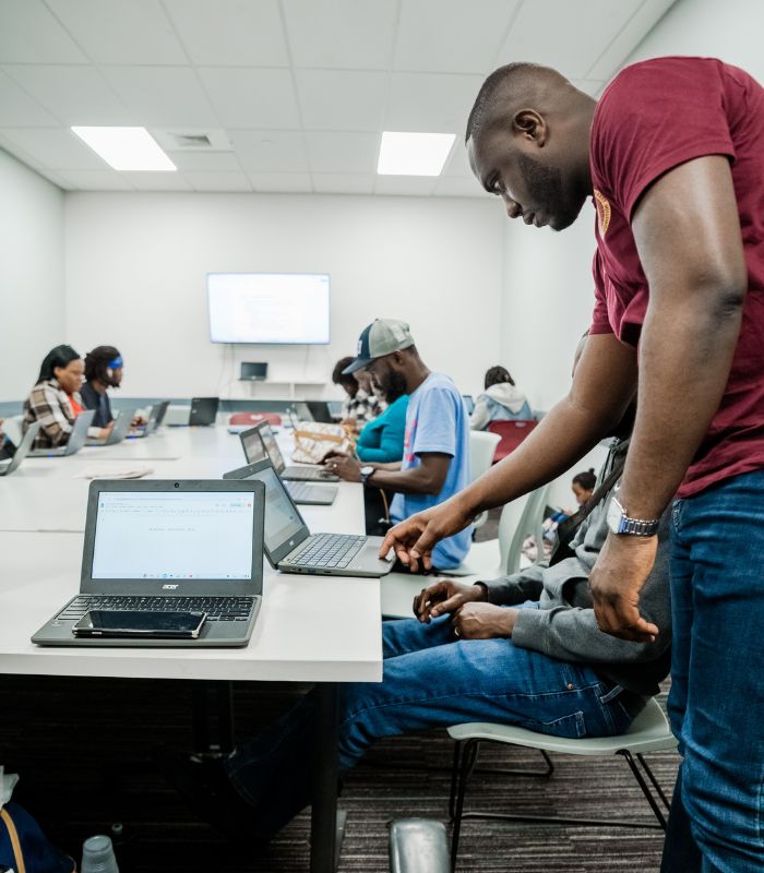 BRICC computer training day at the YMCA. A classroom or training room with several people seated at tables working on laptops. A man in a maroon shirt stands beside a participant, offering assistance. The room is brightly lit with ceiling lights, and a large screen at the front displays presentation content. The atmosphere suggests a hands-on workshop or computer-based learning session.