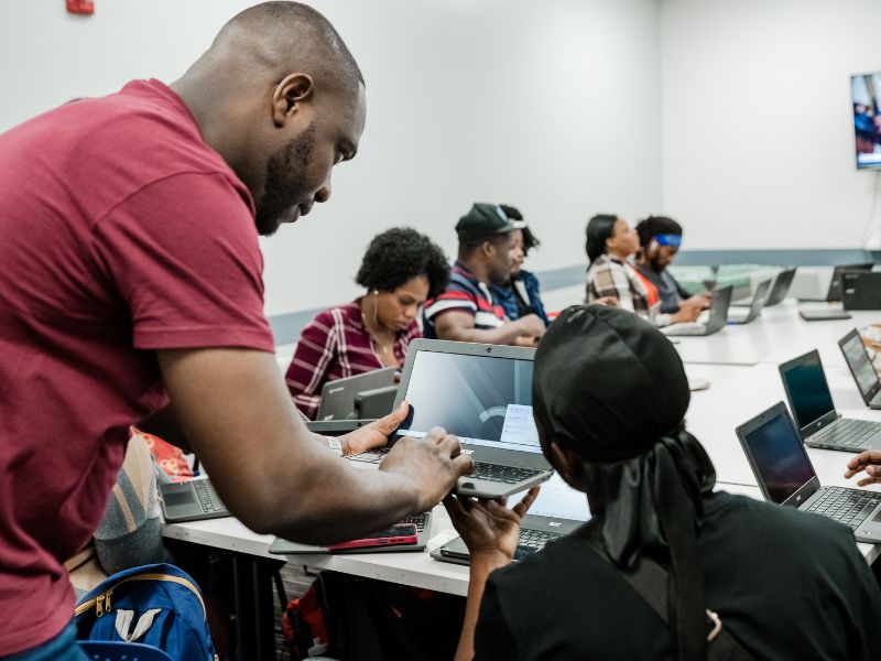 BRICC computer training day at the YMCA. A classroom or training room with several people seated at tables working on laptops. A man in a maroon shirt stands beside a participant, offering assistance. The room is brightly lit with ceiling lights, and a large screen at the front displays presentation content. The atmosphere suggests a hands-on workshop or computer-based learning session.