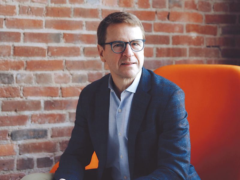 Photo of Dwight Poler sitting on an orange chair in front of a brick wall. He is wearing a navy jacket, blue button down, and tan pants.
