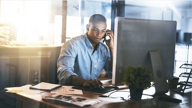 A man of color sitting at a desk in front of a computer. He is holding a cell phone up to his ear.