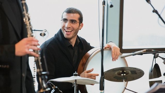 Guy Mendilow playing the drums, wearing a black collared shirt. A man is to the left of him playing a wind instrument.