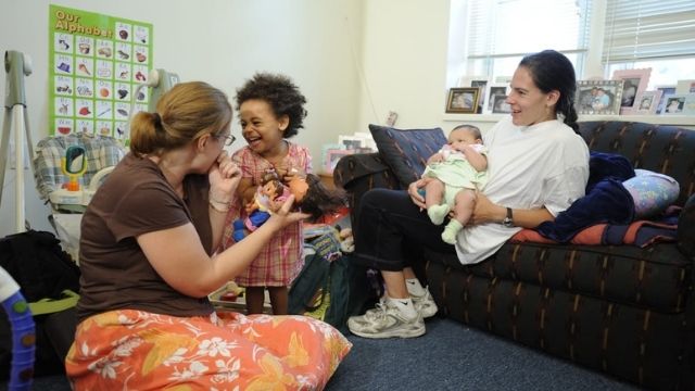 A woman sitting on the floor with a young child - they are smiling at the each other. Another woman is sitting on a couch to the right, smiling at them.