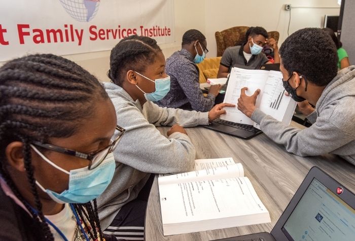 Students wearing masks, sitting at a large table with books and laptops, studying. 