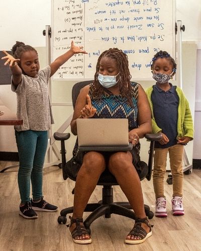 A teacher sitting in a desk chair with two young children on either side of her and white board with lyrics on it in the background. The children look like they are singing with her.