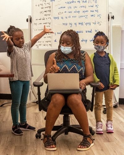 A teacher sitting in a desk chair with two young children on either side of her and white board with lyrics on it in the background. The children look like they are singing with her.