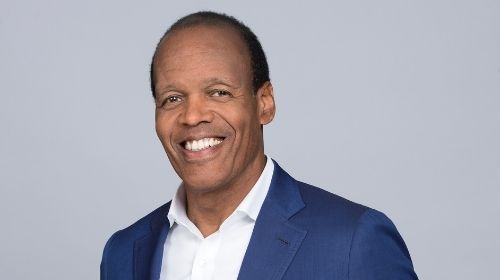 Lee Pelton wearing a blue blazer and white collared shirt. The background is light gray.