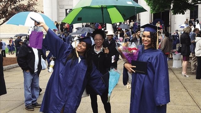 Three college graduates wearing indigo robes standing next to each other, smiling. The middle on is holding an umbrella.