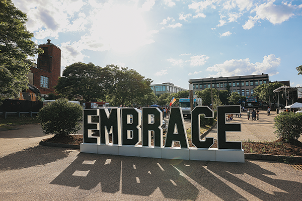 A large cement sign reads "EMBRACE." It is placed in a courtyard surrounded by trees and buildings.