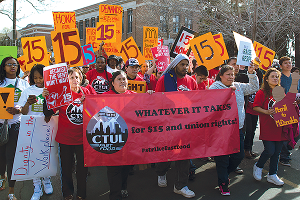 A group of protestors in Kansas City march holding signs that say 15. In front is a large banner that reads Whatever it Takes for $15 and union rights