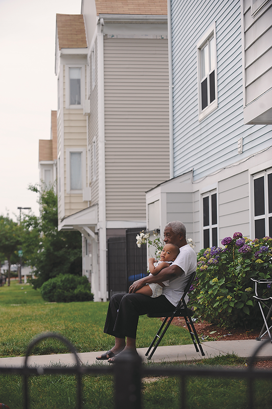 An elderly black man sits on a folding chair on the front walkway to a blue house, he has a toddler in his lap.