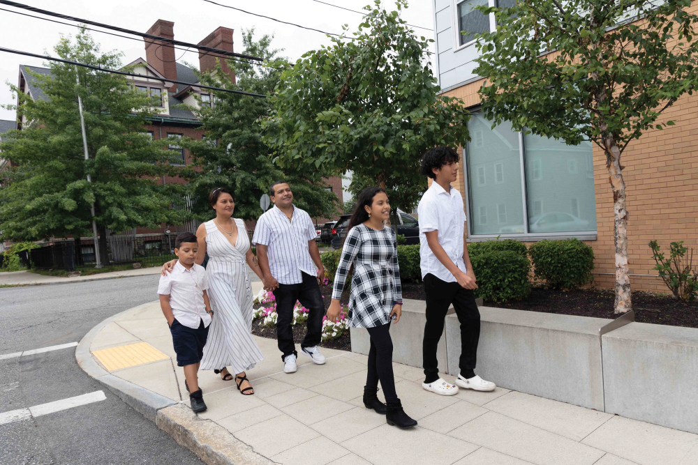 A family of five people walks along a sidewalk near housing