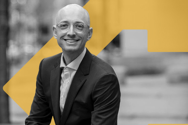 A black and white photo of Luc Schuster. He is a white man, he wears a suit and sits on a stone bench outside. Layered behind Luc is the orange TBF arrow icon.
