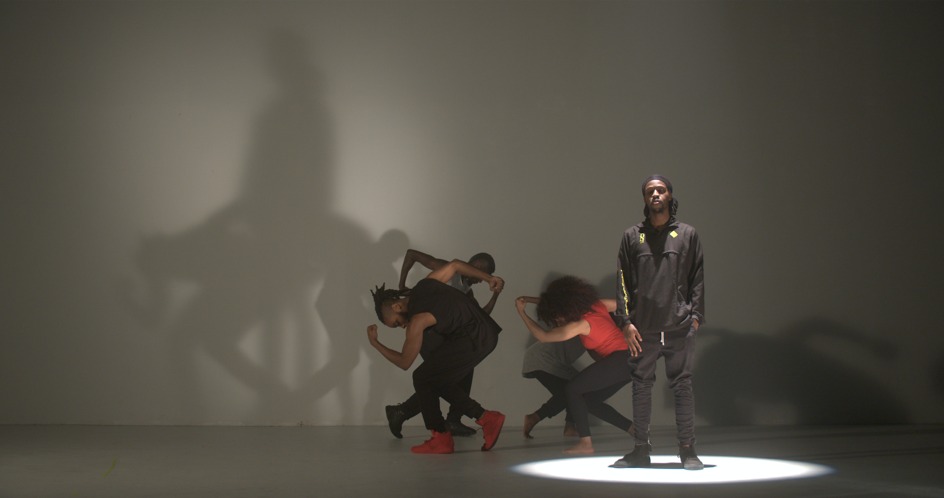 A picture from a beheard.world performance. Five dancers are on a stage. Four are crouched in formation behind one who is standing. All five are off to the right side of the photo. A spotlight is shining on them.