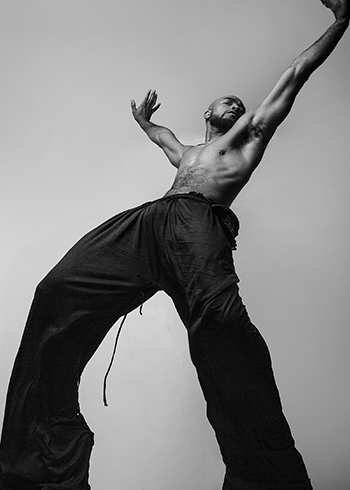 A black and white photo taken at the perspective of the ground shows Anthony Burrell dancing with his legs spread and arms held wide above his head.