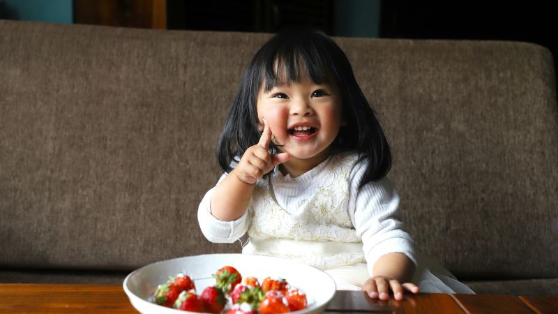 A toddler sits with a bowl of strawberries in front of her, she smiles and points at the camera
