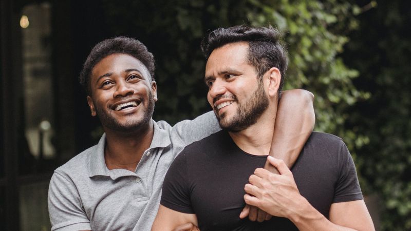 Two men walk down a street with their arms around each other and smiling