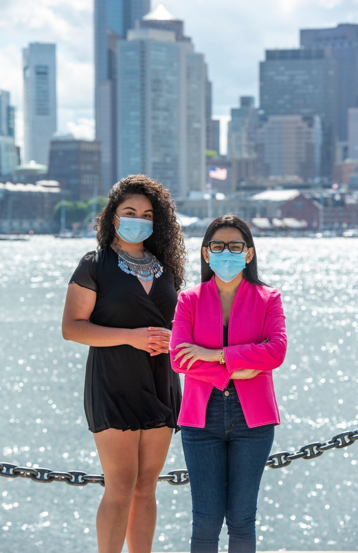 Alexa Cuellar and Patricia Montes standing next to each other. They're both wearing blue face masks. Alexa is wearing a short-sleeved black romper and a turquoise necklace. Patricia is wearing a pink blazer and blue jeans. Behind them is the Boston Seaport and city buildings.