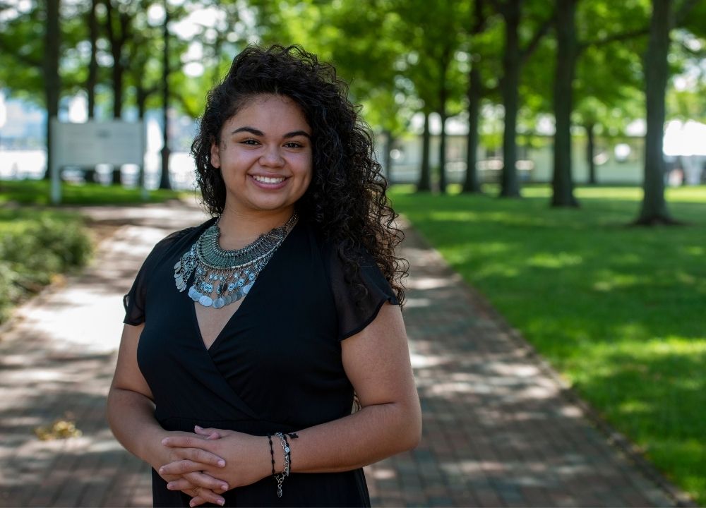 Alexa Cuellar standing outside, a brick walkway trailing behind her. Green trees and grass on either side of the walkway. Alexa is wearing a black dress and a large blue beaded necklace. Her dark, curly hair is swept over her left shoulder.
