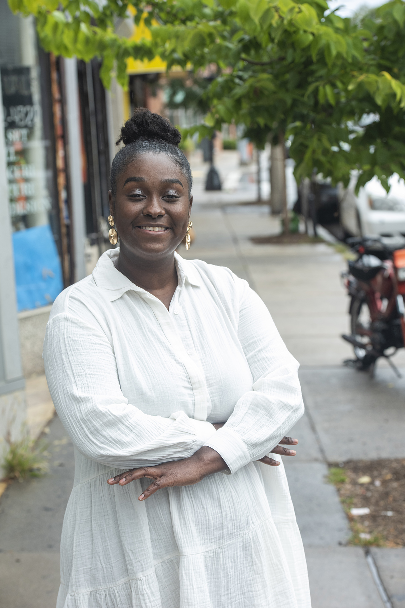 Ameina Mosley standing on a sidewalk, the sidewalk trailing behind here. Storefront windows are seen behind here and to the left. She's smiling, ams crossed, her dark hair in a high bun and she's wearing a white long sleeved dress.