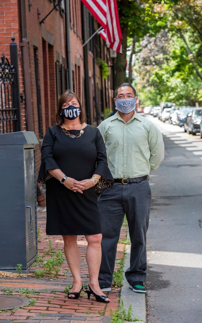 Amy O'Leary and Cliff Kwong standing on a brick sidewalk in Beacon Hill. Trailing behind them are brick buildings on the left, the brick sidewalk, and a paved road to the right. Green trees in the distance and an American flag hanging at the above them. It's a sunny day.