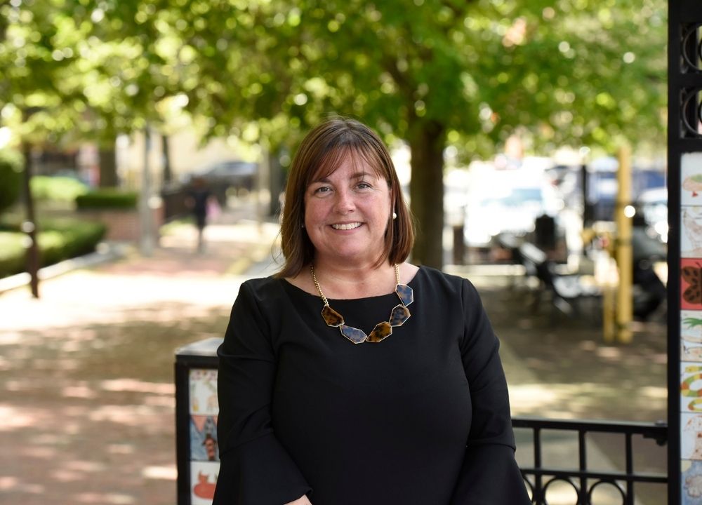 Amy O'Leary standing outside, a brick sidewalk trailing behind her along with green trees. She's wearing a black dress and a stone necklace, smiling. She has short brown hair and bangs. It's a sunny day.