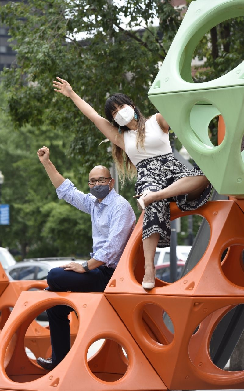 Ben Hires and Yanyi Weng sitting on an orange and green jungle gym somewhere in Chinatown. Both are wearing cloth face masks. Ben has his a fist in the air, and Yanyi has one hand in the air. Green trees in the background.