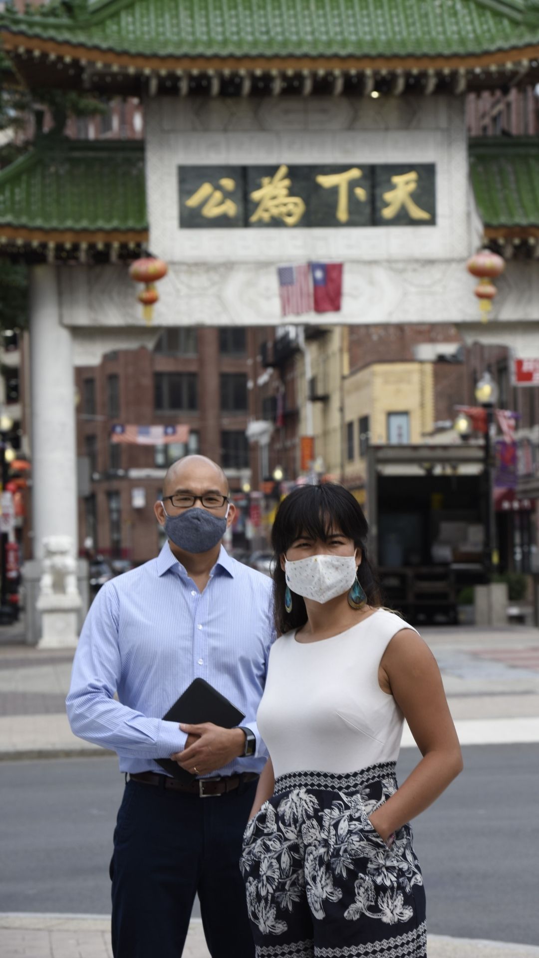 Ben Hires and Yanyi Weng standing next to each other in Chinatown. They're standing in front of and across the street from a green and white Chinese architecture-styled structure with a green and gold sign with Chinese characters on it. Behind the structure are city buildings. Yanyi is wearing a black and white sleeveless dress. Ben is wearing a light blue collared shirt and black pants.
