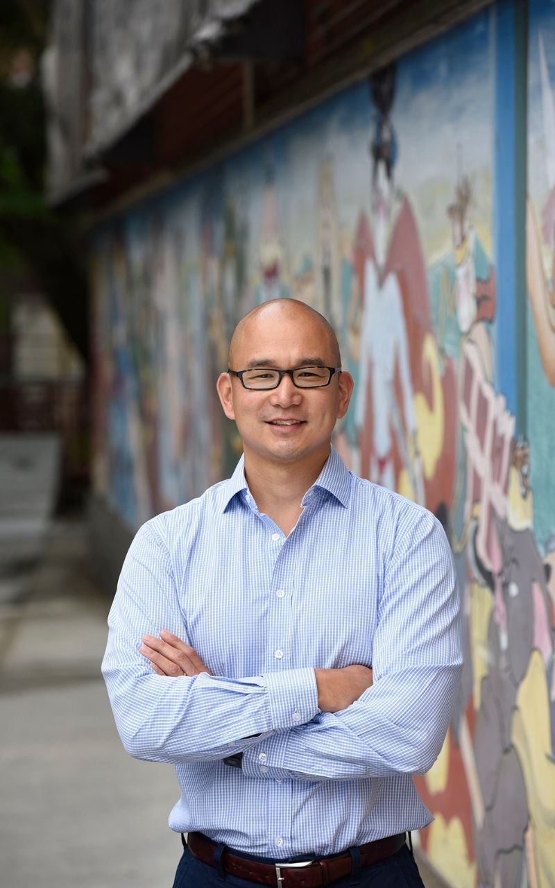 Ben Hires standing outside on a concrete sidewalk, arms crossed and smiling. He's wearing a light blue, long-sleeved, collared shirt, and dark-rimmed glasses. A large wall covered in colorful artwork trails behind him.