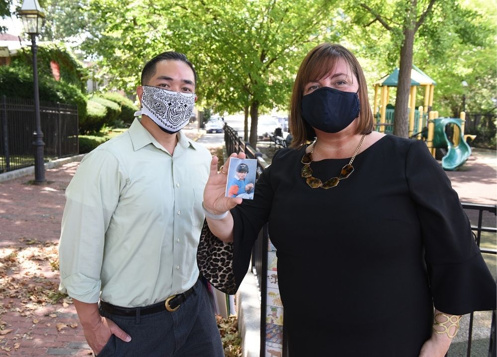 Cliff Kwong and Amy O'Leary standing next to each other wearing masks. Amy is holding up a photo of a young boy in a baseball uniform. A brick sidewalk trails behind them, with green trees in the background. It's a sunny day.