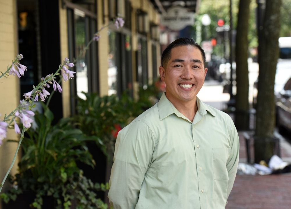 Cliff Kwong standing with his arms behind their back, smiling. He's wearing a long-sleeved, collared, light green shirt. He's standing on a brick sidewalks that trails behind him, along with a yellow building with storefronts and green shrubs on the left. Small purple flowers are peaking into the left side of the image.