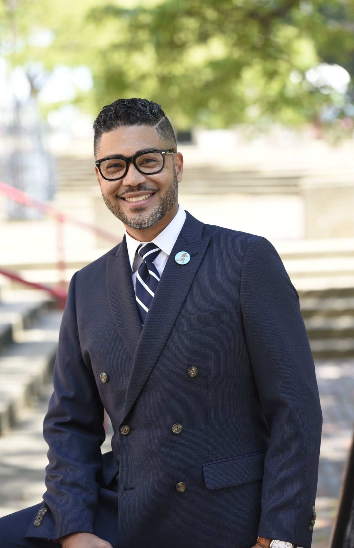 Imari Paris Jeffries smiling, standing with one arm resting on his knee, wearing a dark blue suit with gold buttons on the front, a white collared shirt and blue and white striped tie. He has short dark hair and dark rimmed glasses. Behind him are concrete stairs with a red railing, a brick-laid ground a green trees.