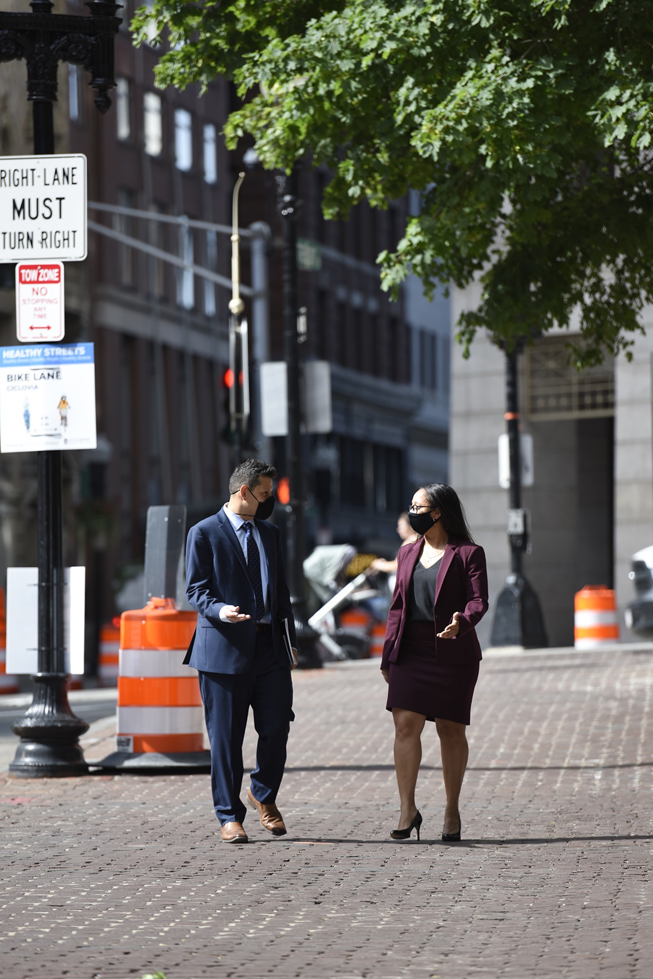 Ivan Espinoza Madrigal and Sophia Hall walking next to each other on a brick sidewalk in the city. They are both wearing masks. He's wearing a dark blue suit and she's wearing a dark purple suit jacket and skirt. They're walking towards the camera and facing each other, gesturing as if having a conversation.