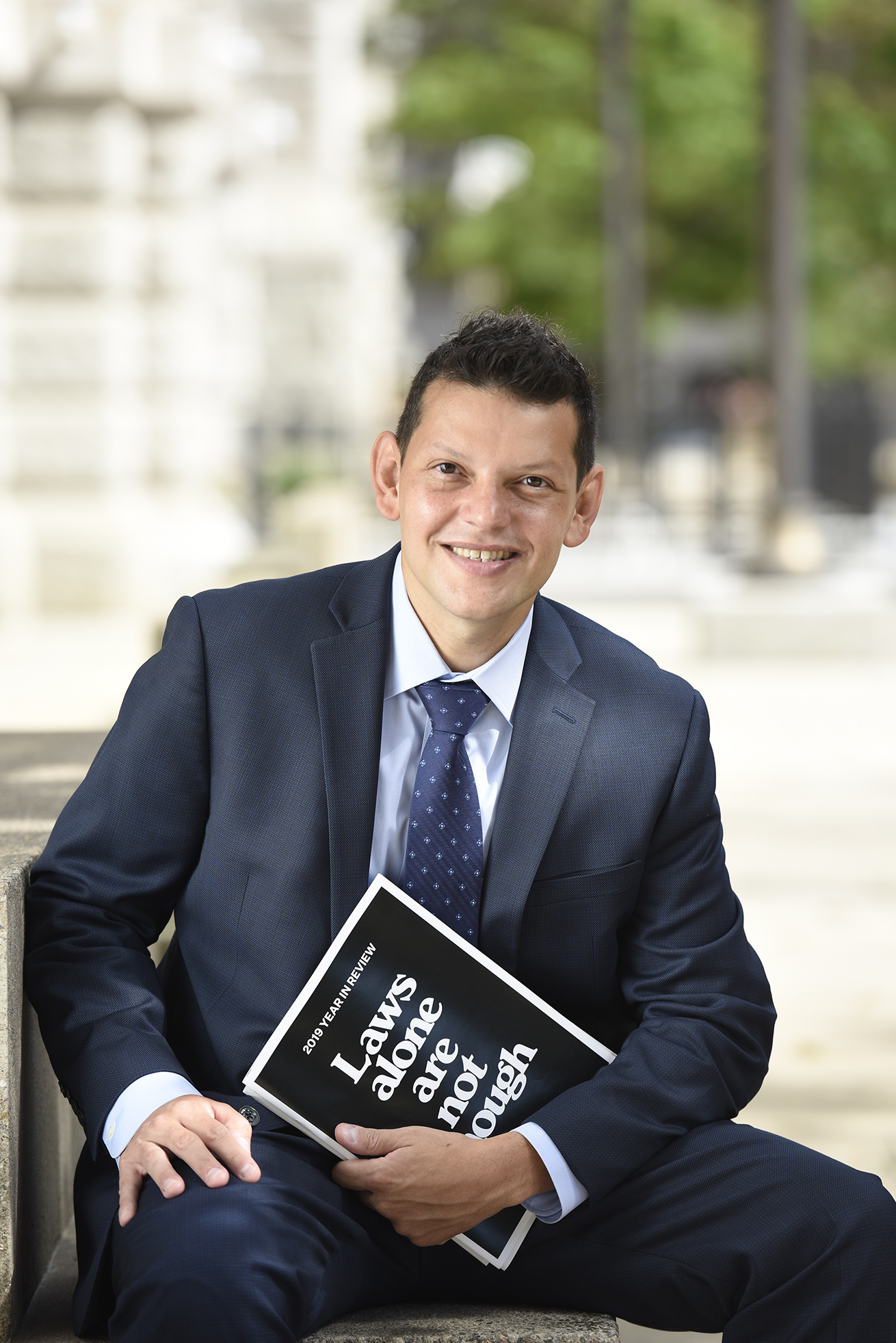 Ivan Espinoza Madrigal sitting on concrete stairs, one hand on one knee and one elbow on the other knee. He's holding a black book that says "Laws along are not enough" i white text on the front. He's smiling and wearing a dark blue suit. A Concrete building and sidewalk trails off behind him, green trees in the background. It's a sunny day.