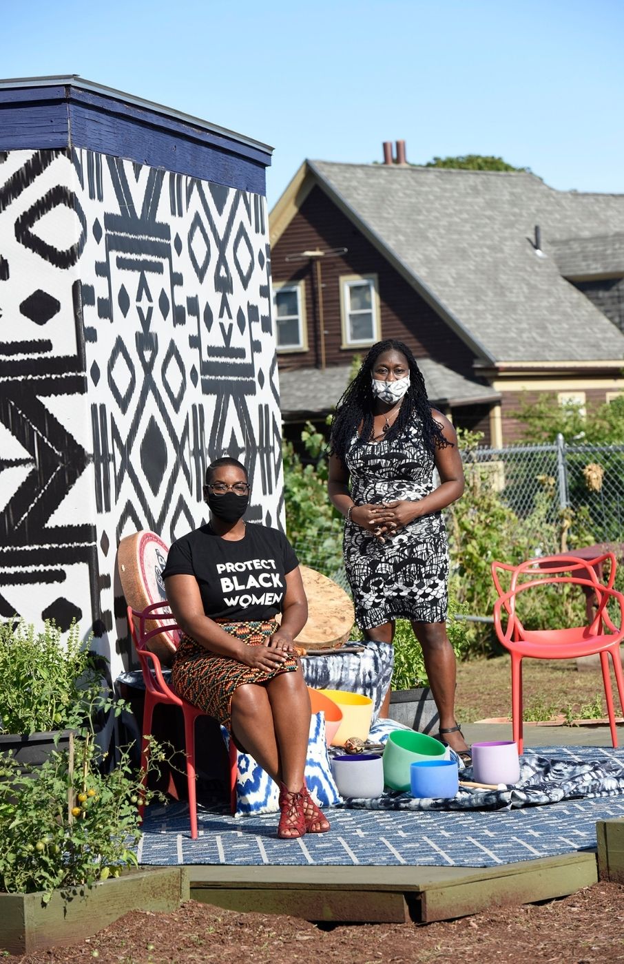 Jennifer Mompoint and Jhana Senxian at The Guild. Jennifer is sitting in an organe metal chair to the left of Jhana, who is standing. Both are on a blue and white patterned wooden platform. Between them are buckets of paint, each one color from the rainbow. Behind them and to the left is a cube structure adorned with a black and white pattern. In the background are bushes, a dirt covered ground, a brown house and blue sky.