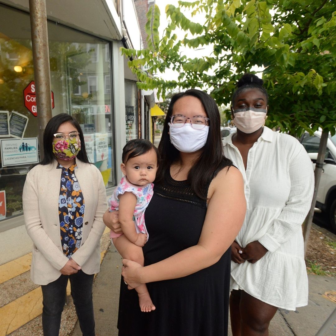 Goribel Rivas, Lily Huang and Ameina Mosely sending next to each other on a sidewalk, the Jobs with Justice facade to their right. Green trees behind them. Lily is holding her baby.